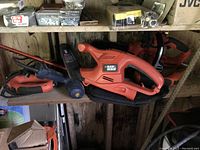 Photo of two corded electric Black & Decker garden power tools on a wooden shelf. Visible are electric hedge clippers and an electric chainsaw with orange and black color scheme.