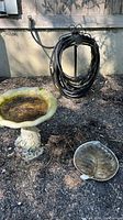 Outdoor photo of all three items on the ground beside a building wall and outdoor faucet: cement birdbath, coiled black hose on reel holder, pewter leaf dish.