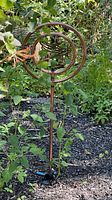 Copper bird wind spinner installed in garden surrounded by greenery and mulch.