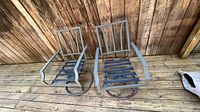 Two extruded aluminum patio chairs with open slat design on seats and backs, visible wear, placed on wooden deck with a wooden wall background