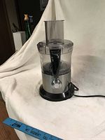Full view of the food processor showing stainless steel body, clear plastic bowl and lid, black handle, and power cord.