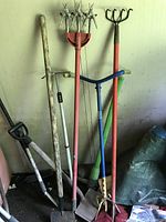Photo showing multiple garden tools leaning against wall including tine aerator, weeding tool, pickaxe, and others