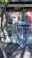 Photo of a large metal bird feeder hanging on a branch-like metal hook attached to a tree outdoors, showing the full bird feeder with a house and garden background.