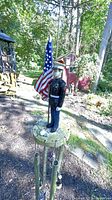 Full view of Marine Corps figurine wind chime with attached US flag outdoors, surrounded by natural setting and structures in background.