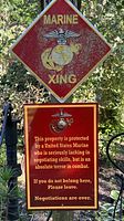 Diamond-shaped Marine XING sign with Marine Corps eagle, globe, and anchor emblem in yellow on red background, weathered and mounted on a post.