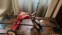 Miniature red tricycle placed on a wooden chest with metal studs, showing the overall condition and design.