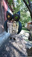 Owl-shaped metal wind chimes with colorful glass eyes hanging on a metal hook outdoors near a wooden wall and foliage.