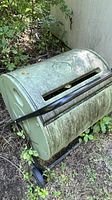Green cylindrical compost tumbler bin on a black metal wheeled frame, showing surface dirt and outdoor wear.