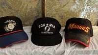 Three baseball caps side by side: left Marine Corps emblem on black with red brim edge, center POW MIA black cap with white embroidery, right black United States Marines cap with red and gold embroidery and red brim edge.