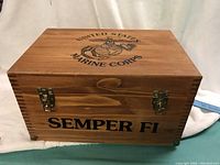 Closed wooden storage box with United States Marine Corps emblem and 'SEMPER FI' text on the front and top lid. Box has two metal clasps for closure.