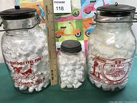 Three vintage glass milk bottles with advertising text in red. They have metal lids and wire handles, placed beside a ruler for scale. Bottles are filled with packing material.