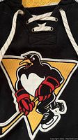 Front logo of Wilkes-Barre/Scranton Penguins penguin holding hockey stick, red gloves, inside a yellow triangle on black jersey fabric with lace-up neckline