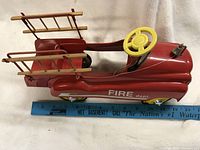 Side angle photo of the red pedal car with attached metal ladder, yellow steering wheel, and Fire Dept. logo visible. Ruler shows length approximately 24 inches.