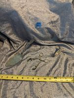 Three antique silver plate items on a grey fabric background with measuring tape: decorative tongs, floral spoon, triangular serving piece.