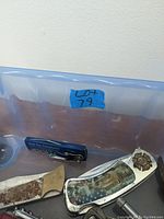 Photo showing close-up of two knives, one with a rusted blade and another with a decorative inlay and badge emblem on the handle.