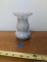 Front view of opaque white glass vase with multi-colored flecks and swirl pattern, positioned on wooden surface with measuring tape showing approx. 10 inches height.