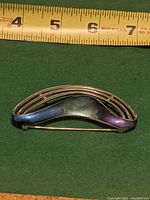 Front view of Mid Century Modernist brooch pin placed against a green background with measuring tape above showing approximately 2.5 inches length, curved dark blue enamel section with silver wire framing.