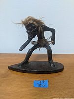 Full view of the primitive Tiki surfer figure standing on an oval wooden base showing the full body and pose.