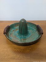 Side view of the signed ceramic vessel showing its turquoise glaze, brown handles, and central juicing cone.