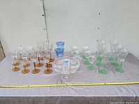 Full view of all glassware including amber, green, and blue stemmed glasses and one clear glass bowl on grey tablecloth.