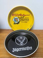 Two round tin beer trays side by side, one yellow Pacifico Clara tray and one black Jägermeister tray, showing logos and general condition.