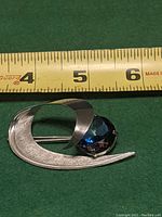 Front view of unsigned modernist brooch with large blue stone beside measuring tape showing size just over 1.5 inches