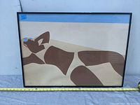 Framed lithograph showing a reclining woman on a beach with sunglasses, minimalist flat shape style, laid horizontally on a ground with measuring tape included for size reference.