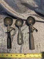 Three vintage ice cream scoops displayed side by side, showing the front with scooping bowls and handles, highlighting two mechanical release handles and one simple scoop.