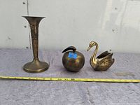 Photo showing three brass items: a tall vase, an apple-shaped container with a blue sticker labeled 'VS', and a swan planter. Items rest on a textured surface with a measuring tape laid horizontally in front.