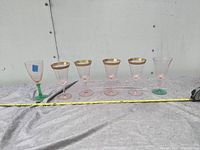 Six pink depression glass goblets arranged in a line on a white lace cloth. Five goblets have gold rims, one has a green bottom. Measuring tape is placed in front for scale.