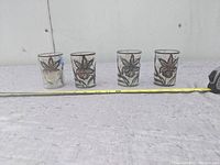 Front view of 4 glass tumblers with silver overlay floral designs lined up next to a measuring tape showing length around 15 inches total.
