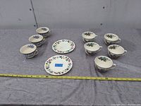 Front view showing all 12 tea cups and 2 floral plates arranged on a fabric surface with measurement tape showing length.