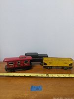 Three vintage toy train cars arranged on a wooden surface with a measuring tape showing approximate 7 to 9 inches length