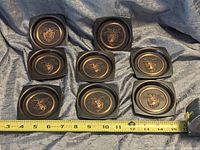 Eight vintage bronze decorative US Military coasters arranged in two rows on fabric background, with measuring tape showing about 7 inches wide.