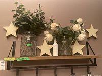 Full view of two decorative glass jars with wire mesh containing greenery and white flowers, surrounded by four wooden stars on a wall shelf.