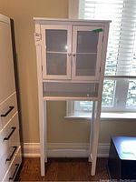 Full view of tall white bathroom cabinet with glass door cupboard and two shelves inside, plus one open shelf below.