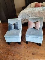 Two light grey upholstered diamond-quilted chairs, each with two velvety cushions in pink and blue, shown side by side on wooden floor near bed.