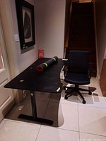 Black office desk with curved wood top, mouse pad cover rolled on top, and black metal legs. Chair positioned next to desk with stairs in background.