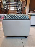 Side view of square ottoman storage cube with white vinyl base and blue geometric patterned cushioned lid on wheels.