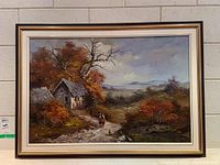 Framed landscape painting showing autumn scenery with a stone cottage and two people on a path