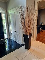 Full view of tall faux plant with thin brown stems in black tapered wood plant stand, placed on light tile floor by black tile entrance.