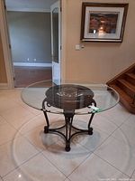 Round glass top table displayed indoors on tiled flooring near stairs and framed artwork, showing full table with glass top and dark metal and wood base.