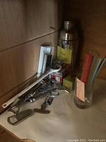 Photo of various bar tools including a metallic cocktail shaker, colored glass stir sticks, metal bottle openers, and other small bar accessories on a wooden shelf.