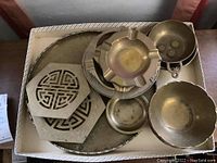View of brass circular tray, brass trivets, ashtray, and multiple brass bowls stacked in a box