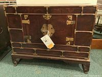 Front and side view of the dark wood campaign chest with brass metal hardware, corner brackets, and decorative lock plate. Chest has a removable wood base with curved legs.