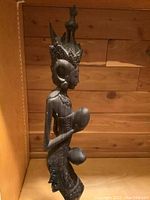 Side view of the carved rosewood statue showing the figure's profile, headdress, and one coconut held near the chest.