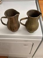 Two copper pitchers next to each other on a white stove top, showing front and side views with handles and spouts visible.