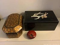 Three lacquer boxes: large rectangular black box with floral inlay, gold octagonal box with black floral pattern, small round red box with floral design.