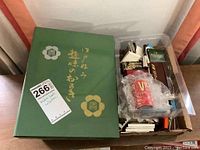 Green box with Japanese writing on the cover beside a plastic container filled with assorted match books.