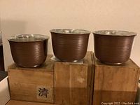 Three spun steel sake waste bowls with brown textured exterior sitting on wooden boxes, showing the ridged detail and polished interiors.
